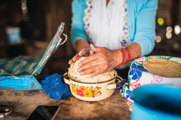 An indigenous Mexican woman wearing a white embroidered blouse and blue sweater is preparing the dough with her hands to make tortillas, a traditional Mexican dish.
