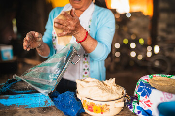 An indigenous Mexican woman wearing a white embroidered blouse and blue sweater, removing the dough from the plastic press to make tortillas, a traditional Mexican dish.
