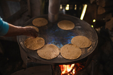 An indigenous Mexican woman wearing a white embroidered blouse and blue sweater, placing flattened dough on the comal to make tortillas, a traditional Mexican dish.
