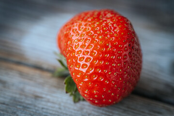Macro image of a deformed ripe strawberry with glistening water droplets, showing bright red skin and green sepals against a rustic wooden background.