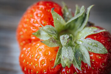 Macro image of a deformed ripe strawberry with glistening water droplets, showing bright red skin and green sepals against a rustic wooden background.