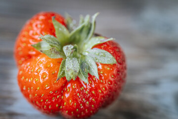 Macro image of a deformed ripe strawberry with glistening water droplets, showing bright red skin and green sepals against a rustic wooden background.