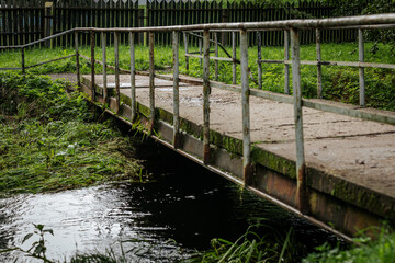 A dilapidated concrete and metal pedestrian bridge crosses the stream, surrounded by overgrown...