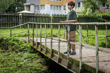 A young boy stands on an old rusted bridge overlooking a grassy stream, with modern apartment...