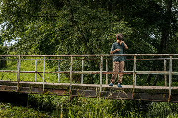 A young boy stands on an old, rusty bridge surrounded by dense foliage, wiping his face in a quiet,...