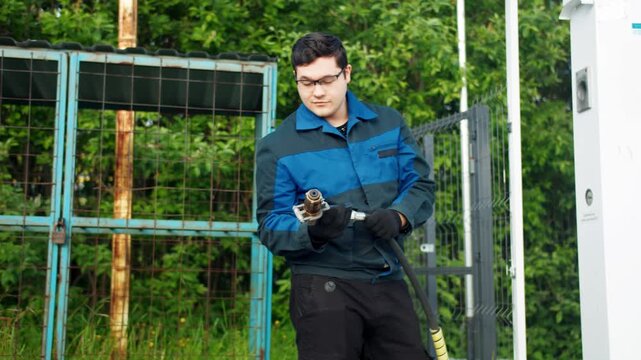 Portrait video of gas station operator in protective uniform picks up a pump gun and smiles at the camera against the backdrop of summer foliage. Confidence of the worker and service personnel