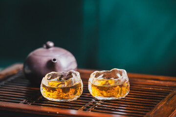 Two glass cups of golden tea on a wooden tray beside a brown teapot, set against a softly blurred...