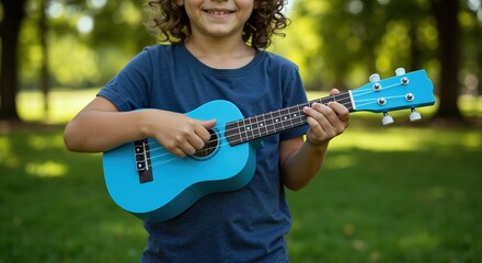 Boy with curly hair playing blue ukulele in navy shirt outdoors. Child learning musical instrument in green park setting. Music education and outdoor learning concept