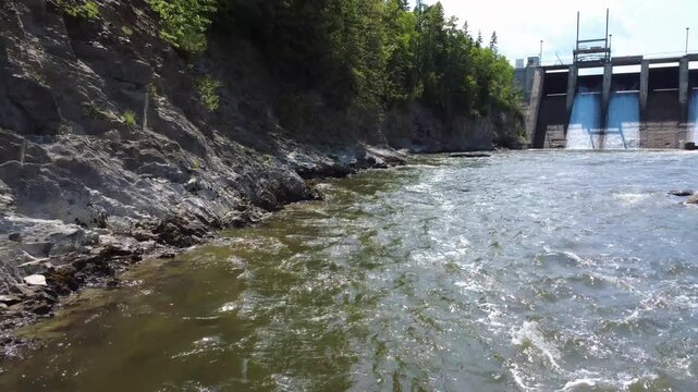 Low aerial footage follows the Mitis River past an eroded forest bank toward the concrete overflow structure of Mitis-2 hydroelectric dam in Price, Quebec, Canada.