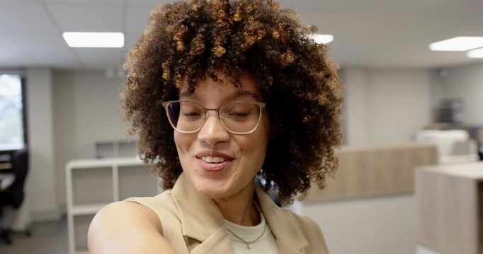Smiling African American woman wearing blazer and clear-frame eyeglasses raising arm in open office