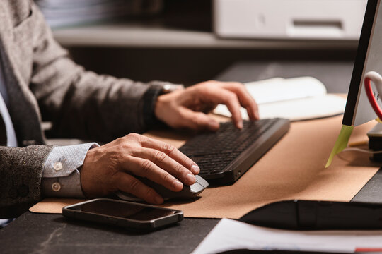Close-up of businessman in suit typing on wireless keyboard at modern office desk. Notebook, mouse, and smartphone visible on workspace.