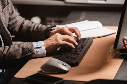 Close-up of businessman in suit typing on wireless keyboard at modern office desk. Notebook, mouse, and smartphone visible on workspace.