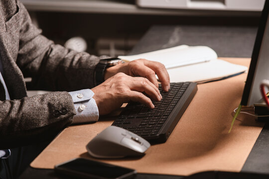Close-up of businessman in suit typing on wireless keyboard at modern office desk. Notebook, mouse, and smartphone visible on workspace.