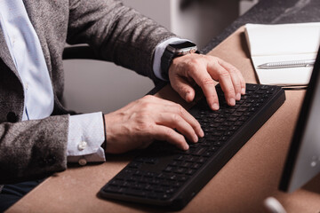 Close-up of businessman in suit typing on wireless keyboard at modern office desk. Notebook, mouse, and smartphone visible on workspace.