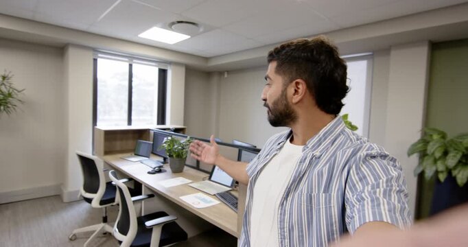 Pointing Indian man showing modular desks in modern coworking space with smartphone, copy space