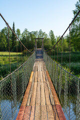 Suspension bridge made of wooden planks stretches across a tranquil river, surrounded by lush...