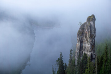 Majestic rock formation rises above a serene river, surrounded by lush evergreen trees and enveloped in a mystical fog, creating a tranquil and enchanting landscape scene