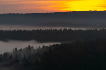 Serene landscape at dawn featuring misty forested hills under a vibrant orange and yellow sky, creating a tranquil atmosphere of nature's beauty and calmness