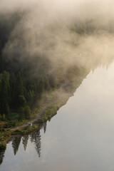 Misty landscape with a winding path along a serene river, surrounded by lush green trees and soft...