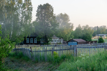 Rustic wooden house surrounded by lush greenery and a wooden fence, with misty atmosphere creating...