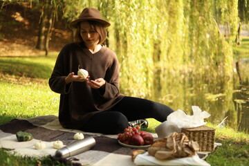 Woman relaxes by the lake, sitting on a picnic blanket surrounded by apples, bread, and warm autumn...