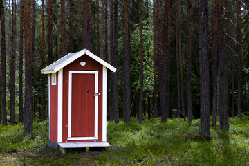 Traditional red wooden toilet outhouse with white edges in the forest with tree trunks and...