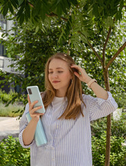 Young woman with long blonde hair is standing outside, smiling, looking at her smartphone and...