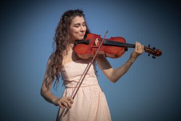A beautiful young female with long hair violinist plays her viola in an outdoor setting, against clear blue sky. Violinist young lady with the alto violin close-up. © Антонина Шатревич
