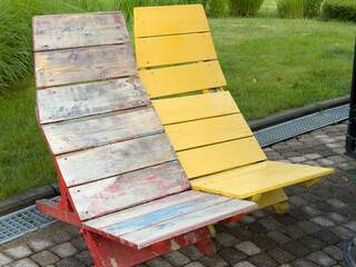 Two colorful wooden lounge chairs are arranged side by side on a paved patio, featuring one with faded red wood and the other in bright yellow, bringing vibrant charm to an outdoor space.
