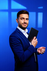 Confident Young Man in a Blue Suit Holding a Tablet Against a Vibrant Blue Background During a Corporate Event
