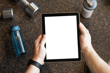 Person holds tablet with white blank screen against gym backdrop with fitness tools, dumbbells,...