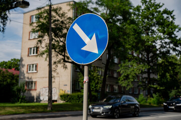 Blue mandatory traffic sign with white arrow pointing down and left, urban street with cars,...