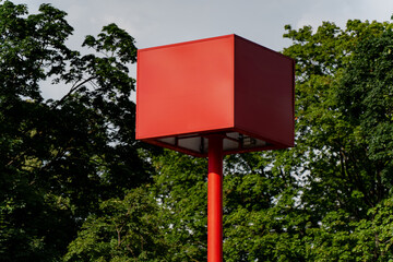 Red square billboard on a tall pole against green trees and cloudy sky, minimalistic urban...
