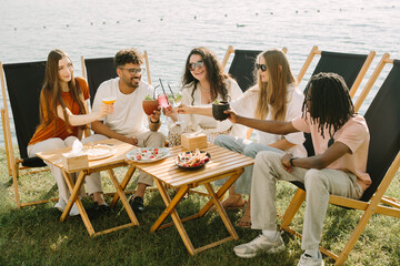 Cheerful friends toasting drinks at lakeside cafe: celebrating summer together
