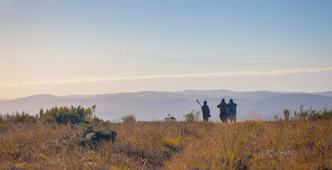 Figures of men with guns and binoculars against the background of the morning hilly landscape.