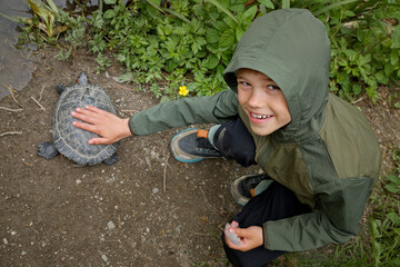 A curious child in a hooded jacket gently touches a turtle on the ground, exploring nature and...