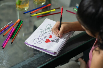 Close-up side view of girl’s hand drawing colorful letters in a notebook on a glass table