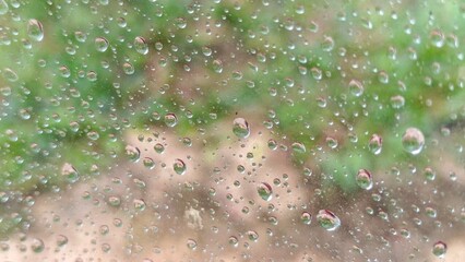 A close-up of water droplets on a windshield surface, with blurry greenery visible in the background.