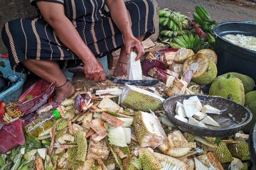 A vendor is cutting jackfruit, surrounded by pieces of the fruit and other produce, preparing it for sale in a vibrant market setting.