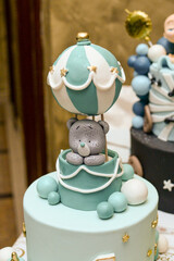 A blue baby birthday cake on top with a teddy bear in a balloon around it is number one, a baby bottle shape and a cube with the letters of the alphabet on it.