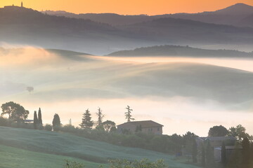 Fototapeta premium Colorful sky of a very foggy sunrise over Val d'Orcia Valley and its podere -farmsteads- among rolling hills. San Quirico d'Orcia-Tuscany-Italy-097