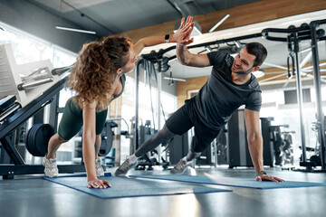 Smiling fit man and woman holding plank position and giving high five during core workout on mats in gym. Team training motivation and healthy lifestyle. Concept: fitness, partnership, energy.