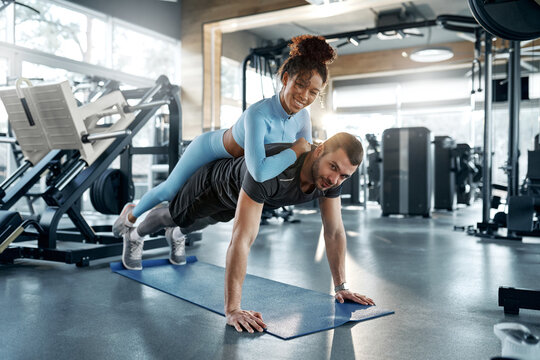 Afroamerican woman balancing on man's back while he does push ups on mat at gym. Concept: fitness, fun training.