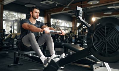 Focused athletic man using a rowing machine during workout at a bright, spacious gym. Concept:...