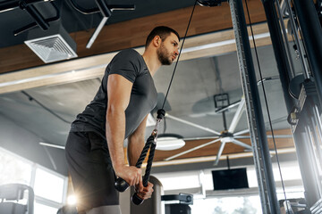 Young athletic man training triceps using rope attachment on cable machine in a modern gym. Intense...