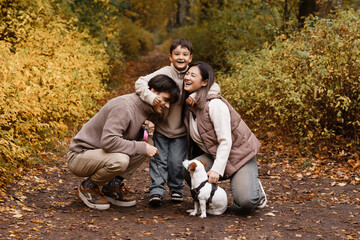Fototapeta premium Front view of young parents and son on a park with small dog, autumn forest background. Concept of family, active lifestyle, pet care, family health, mortgage and family security.