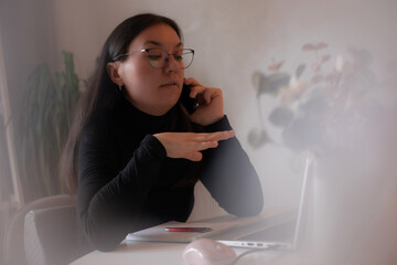 Woman in glasses talking on smartphone while working on laptop at home, gesturing with hand, seated...
