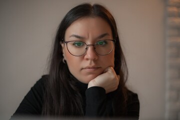 Close-up of a serious woman with glasses resting her chin on her hand, looking at a screen with...