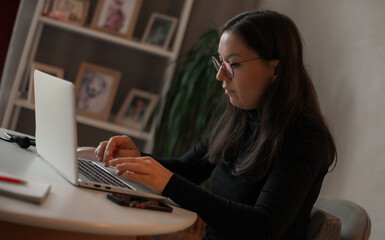 Woman in glasses typing on laptop at home office desk, focused on work, with framed photos and...