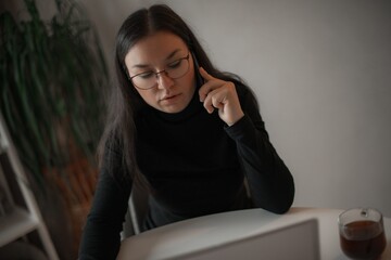 Young woman in glasses speaking on the phone while working on a laptop at home, seated at a desk...